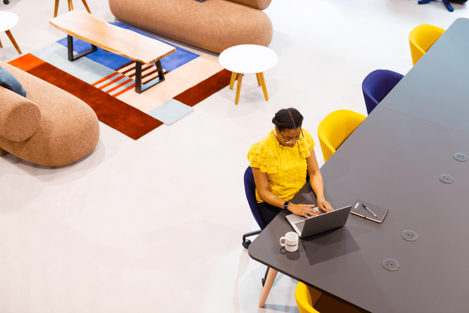 A woman working at laptop at a long table. She is wearing a yellow top. The photo is shot from above.