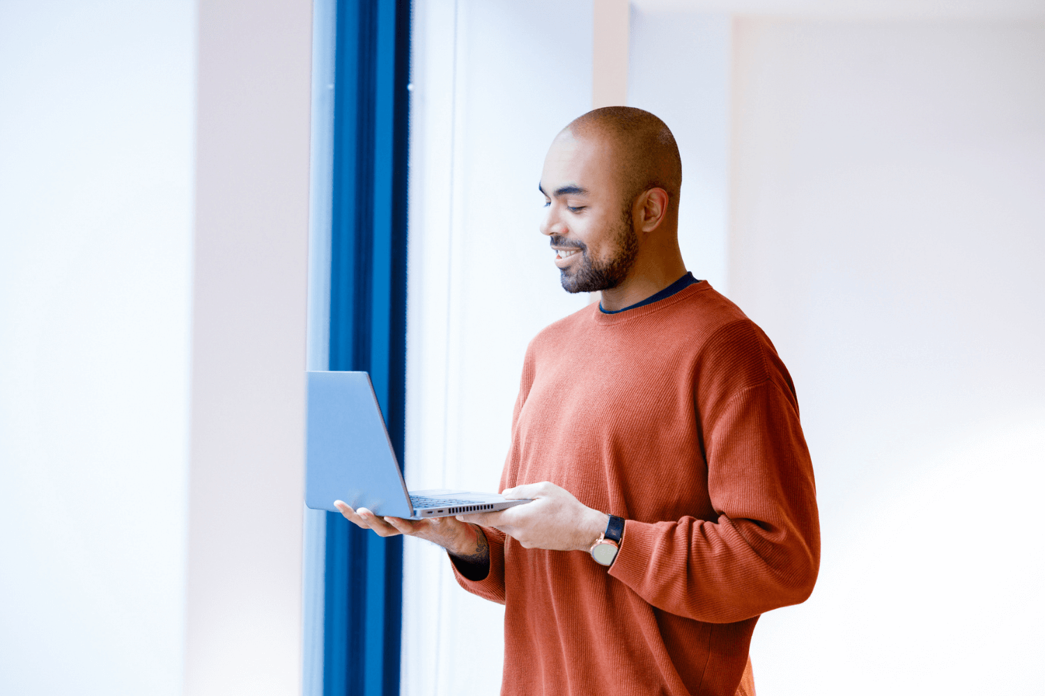 A man in an orange jumper, leaning against the wall and looking at his laptop. 