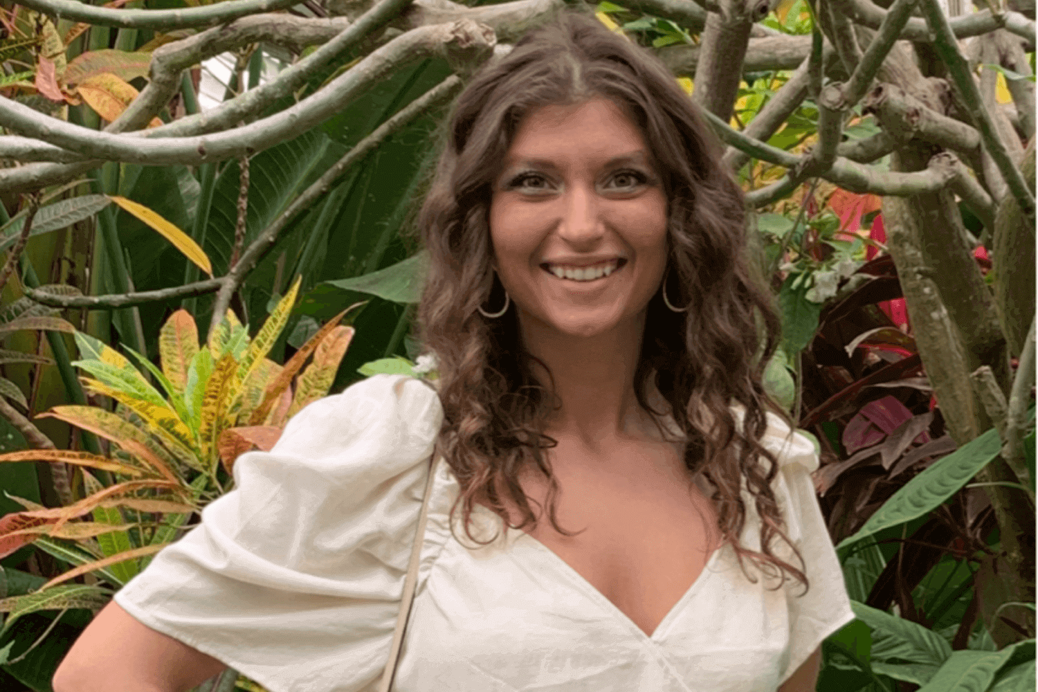 A woman in a white short sleeved top and jeans. She is surrounded by plants.