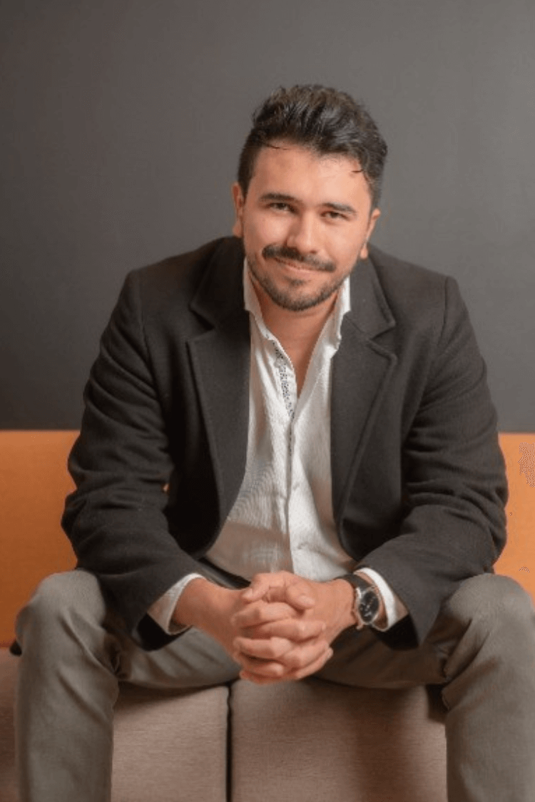 A man sat on an orange and brown sofa. He is wearing grey chinos. a white shirt and a grey jacket. 