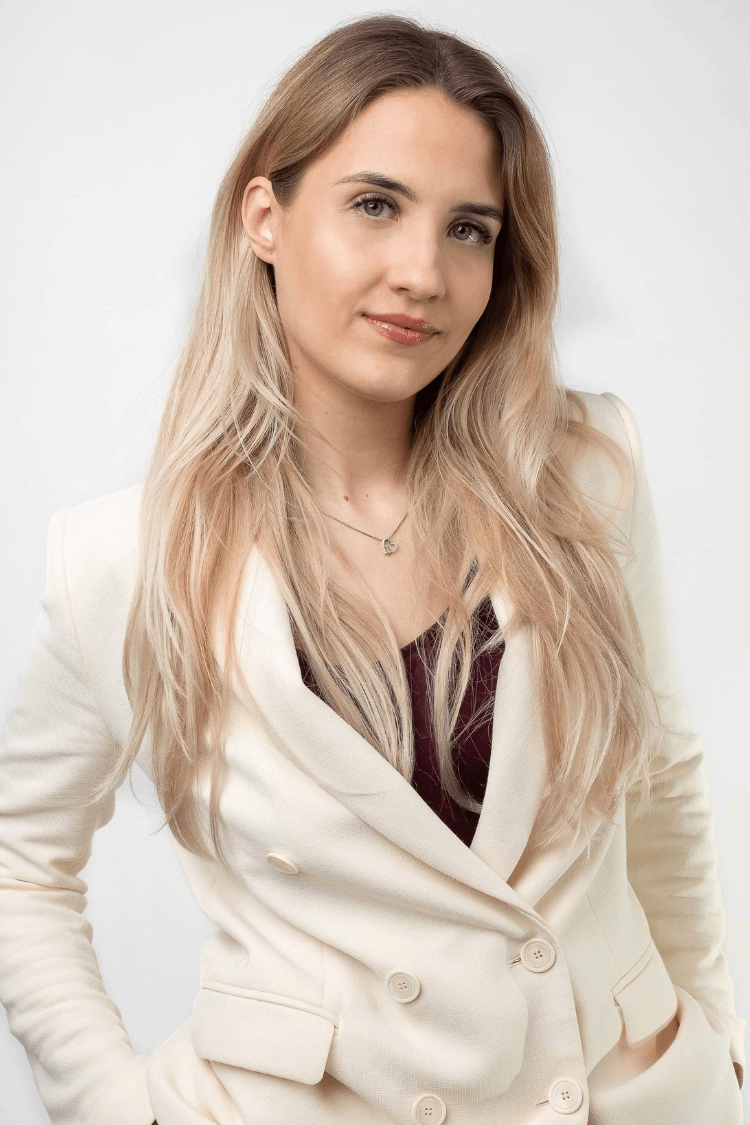 A woman with long blonde hair, wearing a white button up jacket, with a red top underneath. 