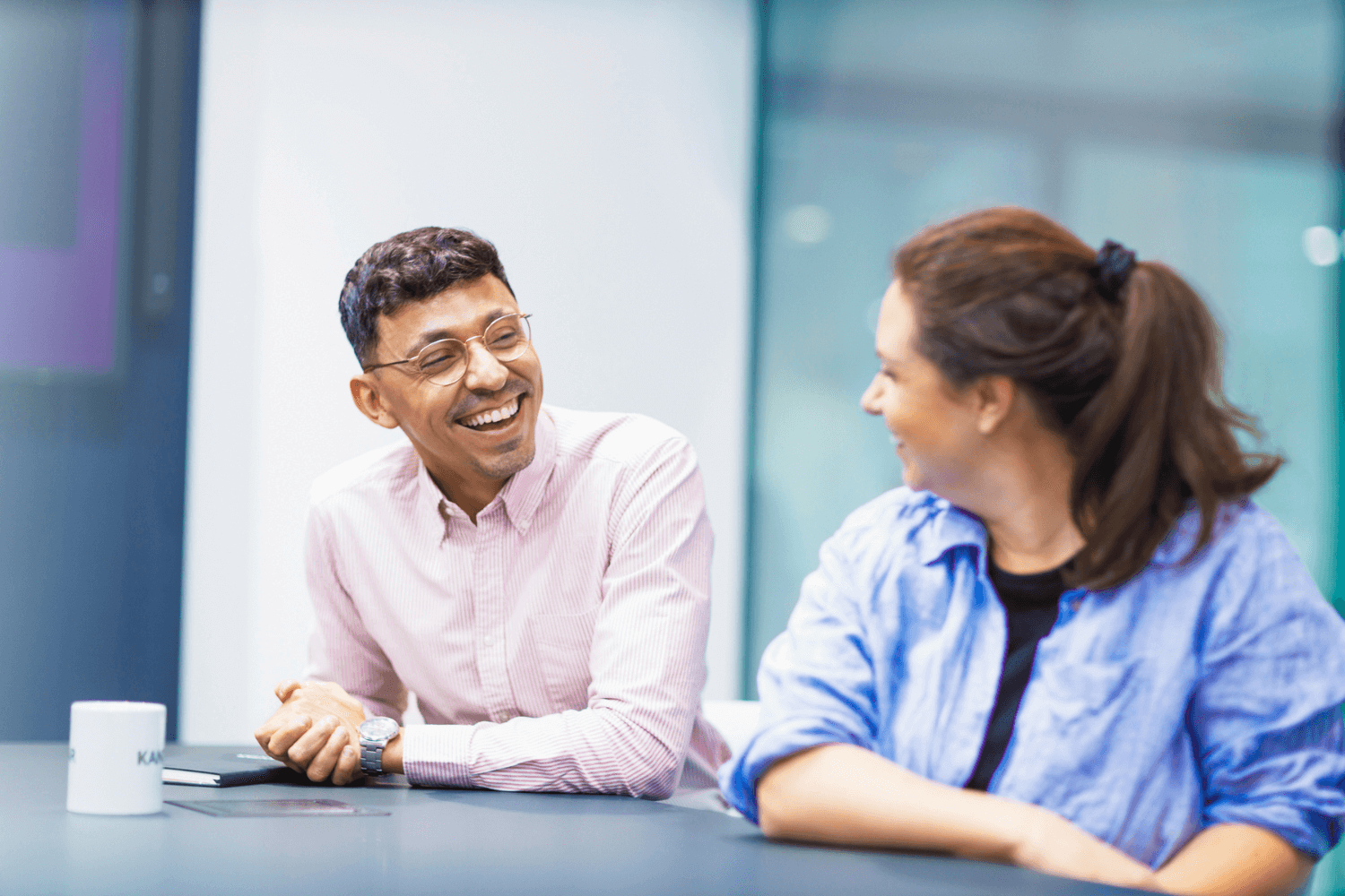 Ein Mann in einem hellrosa gestreiften Hemd und mit Brille sitzt an einem Tisch. Er lächelt die Frau an, die neben ihm sitzt. Sie trägt eine hellblaue Short und hat dunkelbraune Haare, die zu einem Pferdeschwanz gebunden sind. 