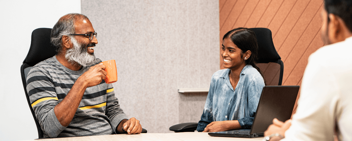 Zwei Kollegen unterhalten sich miteinander. Links ist ein älterer Herr. Er ist Inder, hat einen grauen Bart und trägt einen grauen Pullover mit schwarzen und gelben Streifen. Er trinkt aus einer orangefarbenen Tasse. Er spricht mit einer jüngeren Kollegin. Sie trägt eine blaue Bluse und hat ihre Haare zurückgebunden.