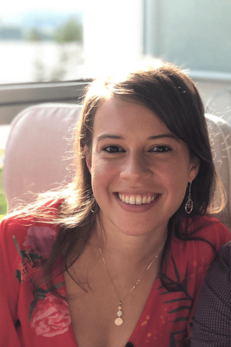 Eine Frau mit dunklem Haar, das etwas länger als schulterlang ist, lächelt in die Kamera und trägt ein rotes Oberteil mit Blumenmuster. 