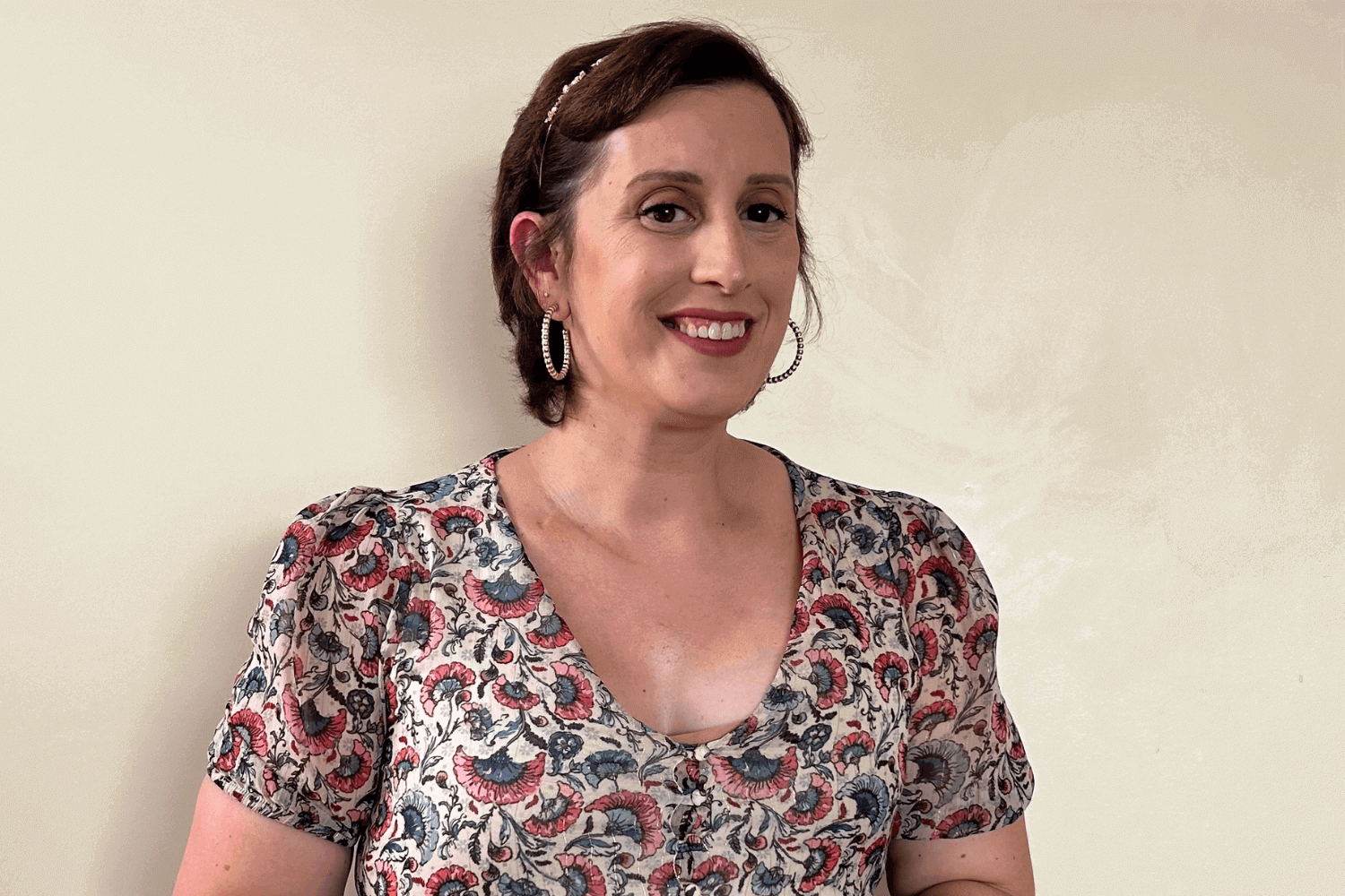 Eine Frau mit kurzen braunen Haaren und goldenen Ohrringen mit Reifen. Sie trägt ein mehrfarbiges, gemustertes Oberteil. 