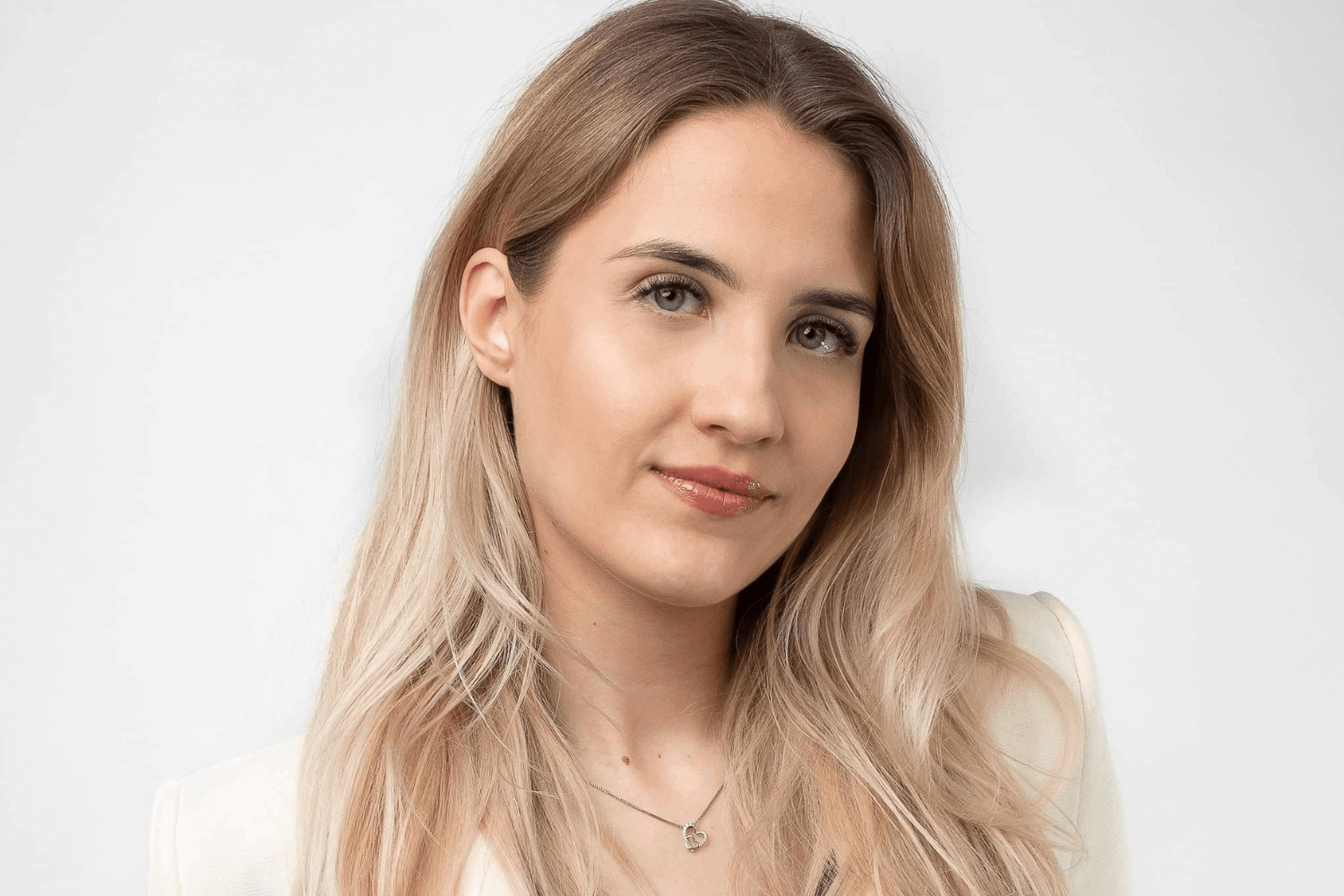 Eine Frau mit langen blonden Haaren, die eine weiße Jacke mit Knöpfen trägt, darunter ein rotes Oberteil. 