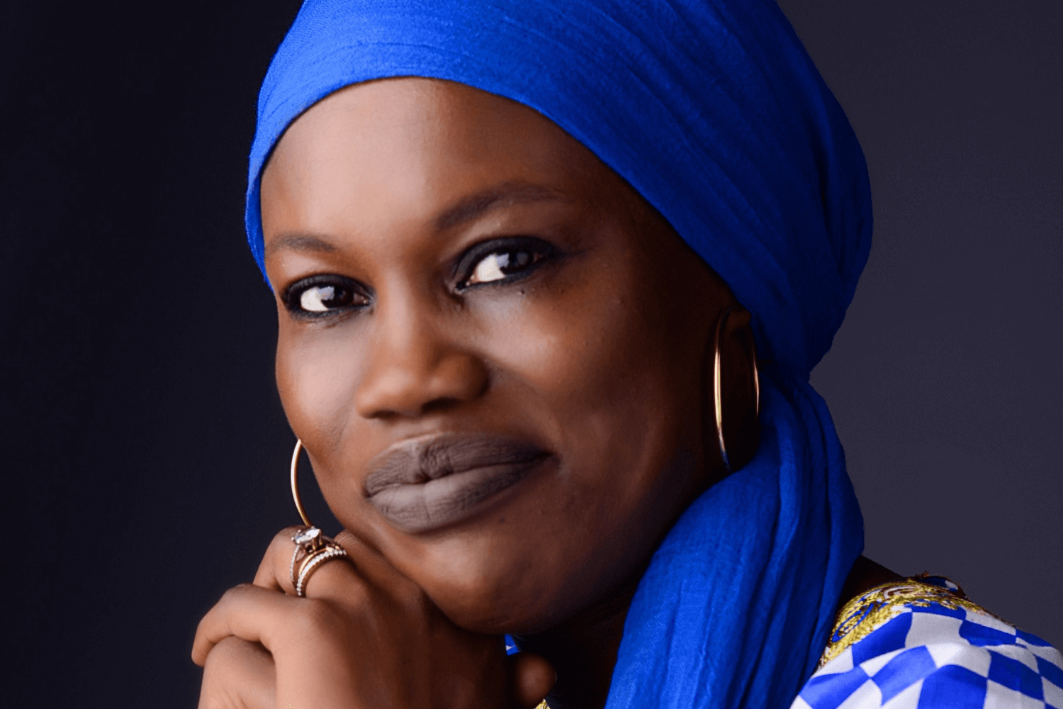 Eine Frau stützt ihren Kopf auf ihre Hände. Sie trägt ein blau-weiß kariertes Hemd und eine königsblaue Kopfbedeckung. 