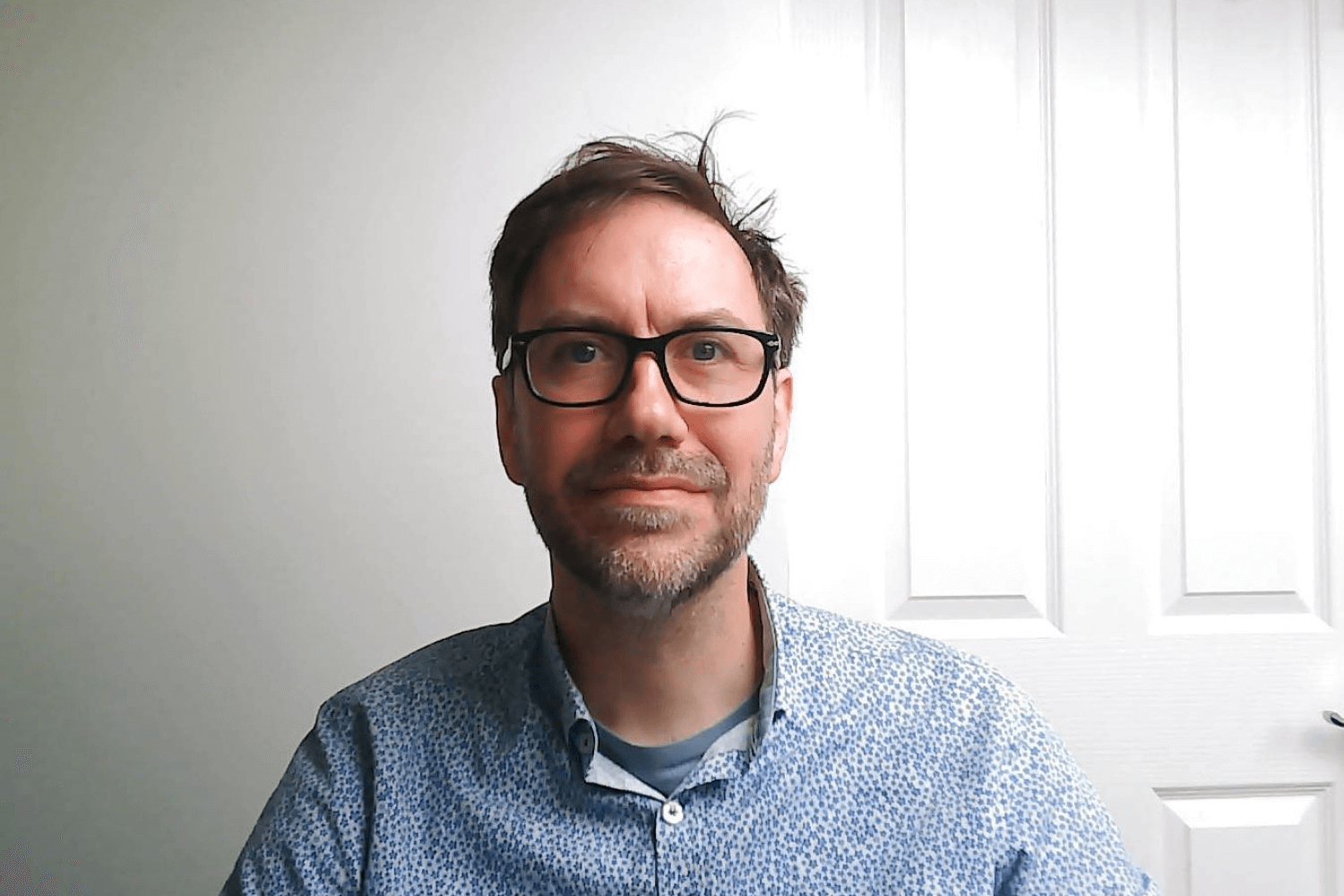 Ein Mann mit einer schwarz umrandeten Brille und einem blauen Hemd schaut in die Kamera. Er hat kurzes braunes Haar. 