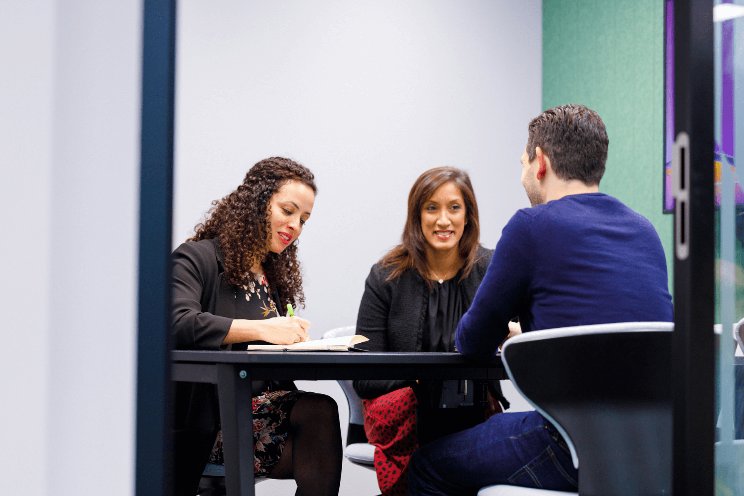 La sala de reuniones es verde, con una mesa en el centro. Hay dos mujeres de frente y un hombre de espaldas a la cámara.