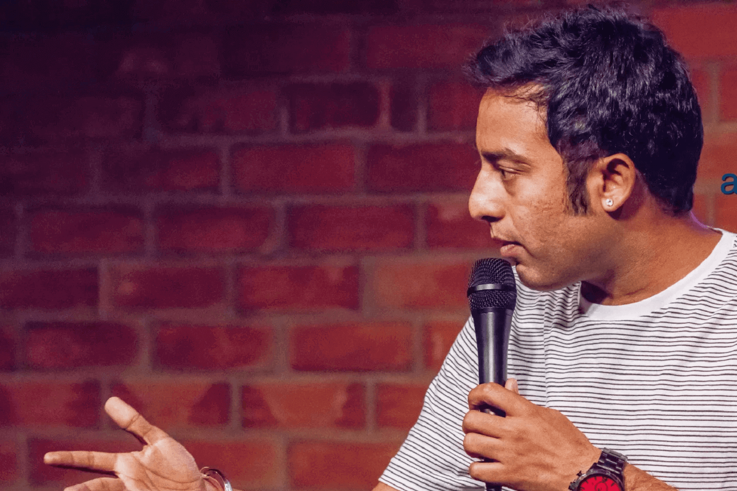 Un hombre en el escenario. Lleva una camiseta de rayas horizontales grises y claras y sostiene un micrófono. 