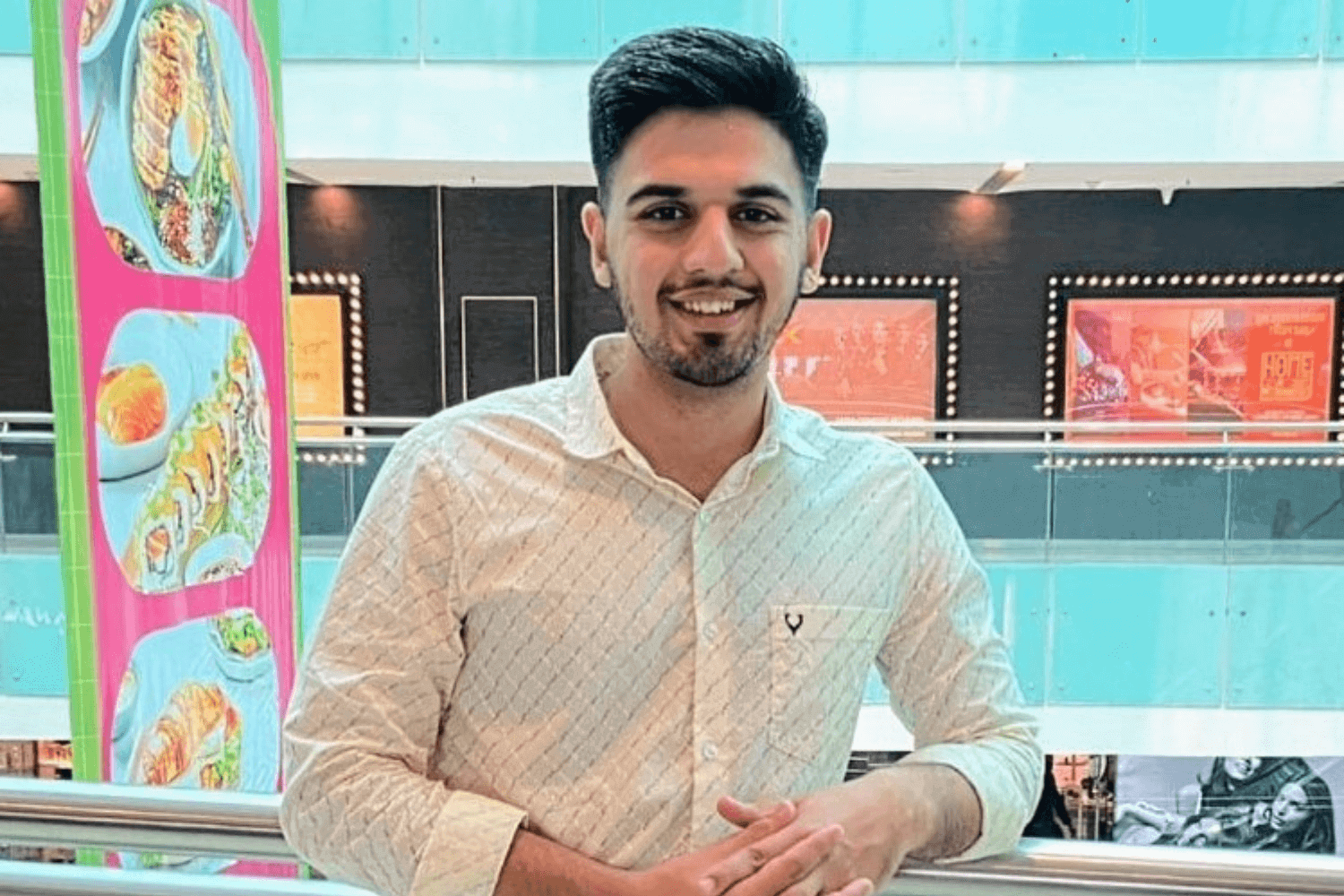 Un hombre con camisa blanca y vaqueros, apoyado en la barandilla de un balcón de un centro comercial. 