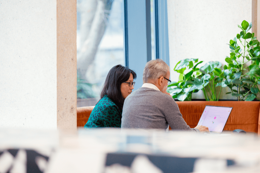 Deux collègues travaillant ensemble à partir d'un ordinateur portable. Elle a des cheveux bruns aux épaules, porte des lunettes et une robe verte. Il porte un pull gris moyen et une chemise blanche.