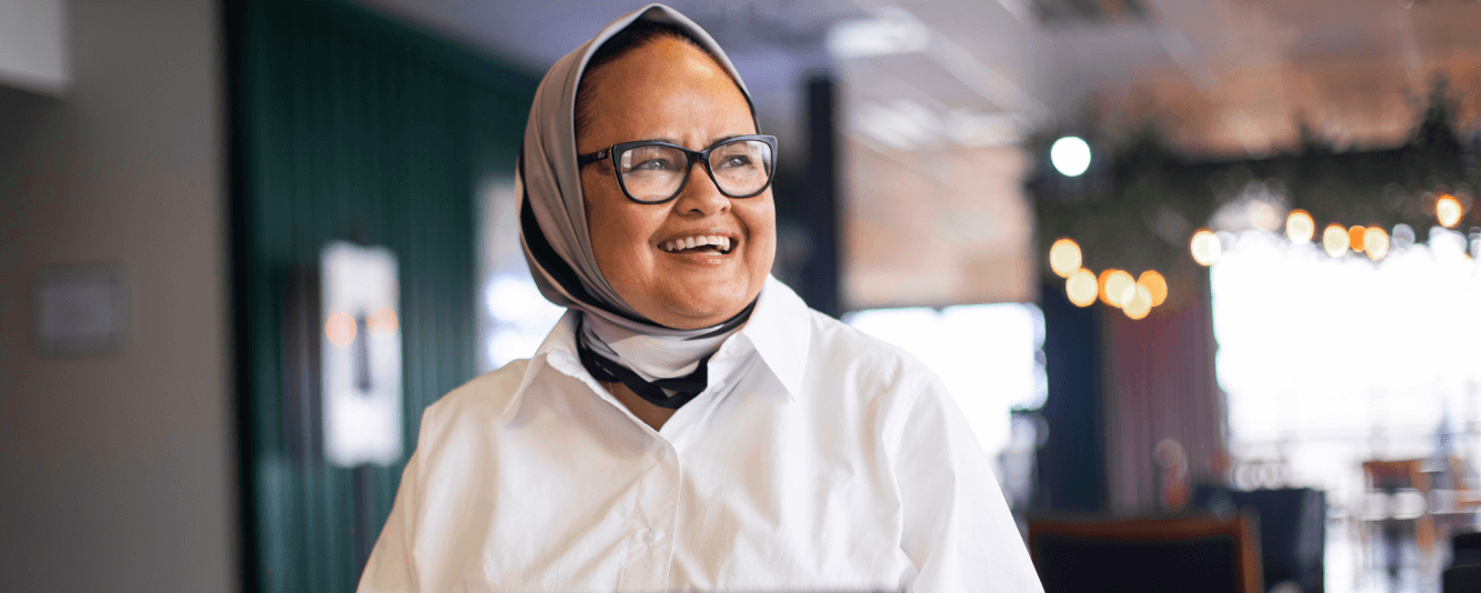 Une femme en chemise blanche sourit vers la droite. Elle porte un foulard, qui est un mélange de couleurs claires et foncées. Elle porte des lunettes.