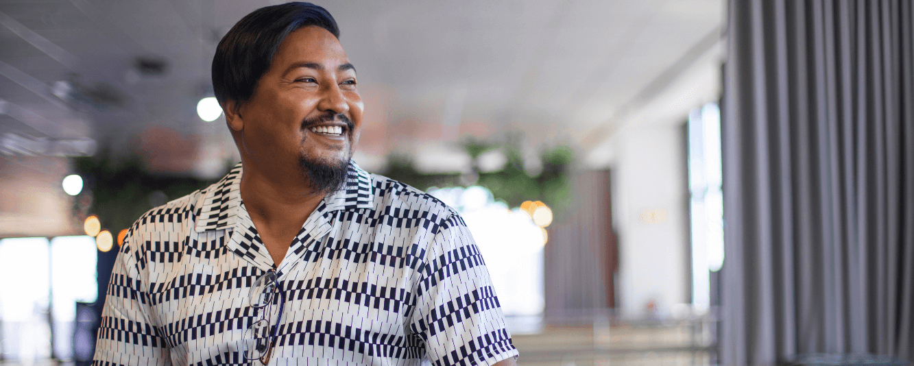 Un homme sourit vers la droite. Il a les cheveux courts et bruns et porte une chemise noire et blanche avec un motif ressemblant à des touches de piano.