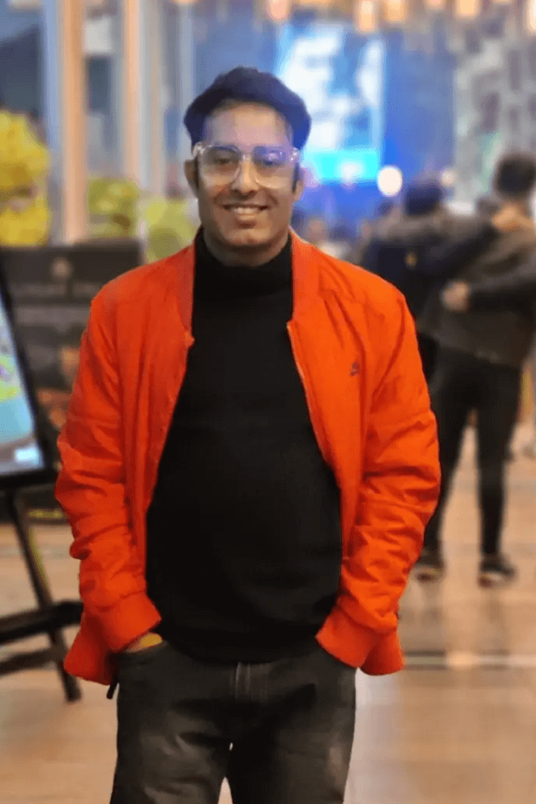 Un homme vêtu d'une veste orange et d'un polo noir, souriant directement à la caméra. Il se trouve à l'extérieur, dans une scène de type café. 