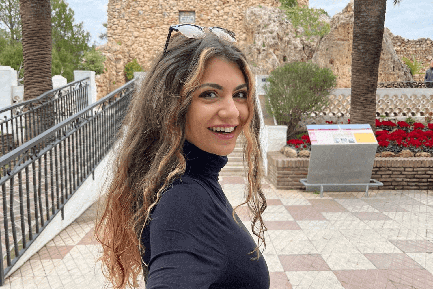 Une jeune femme regarde la caméra. Elle a les cheveux bouclés et des lunettes de soleil sur la tête. Elle porte un polo noir. 