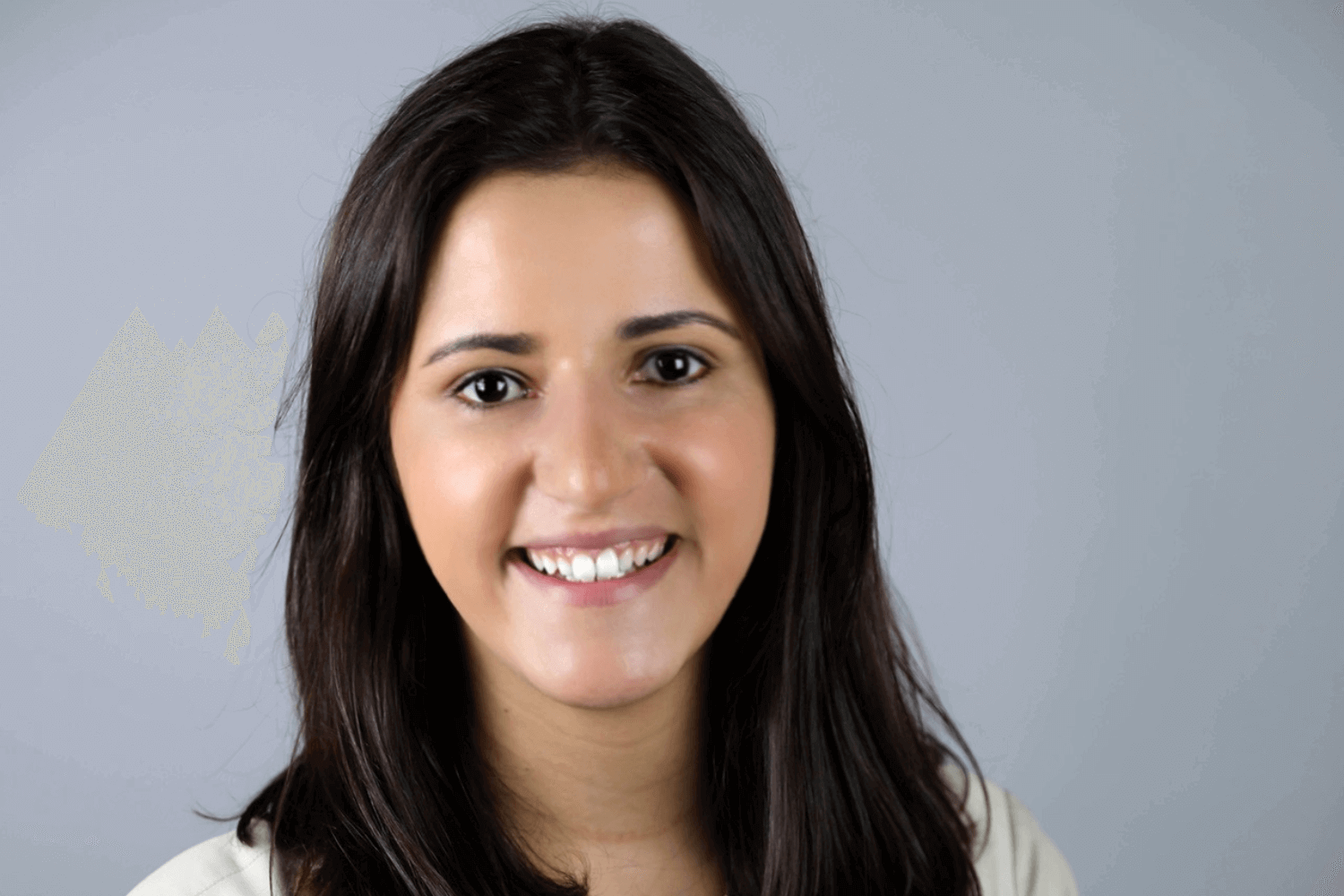 Une femme sourit à l'appareil photo, les bras croisés. Elle porte une veste crème et des cheveux bruns au niveau des épaules. 