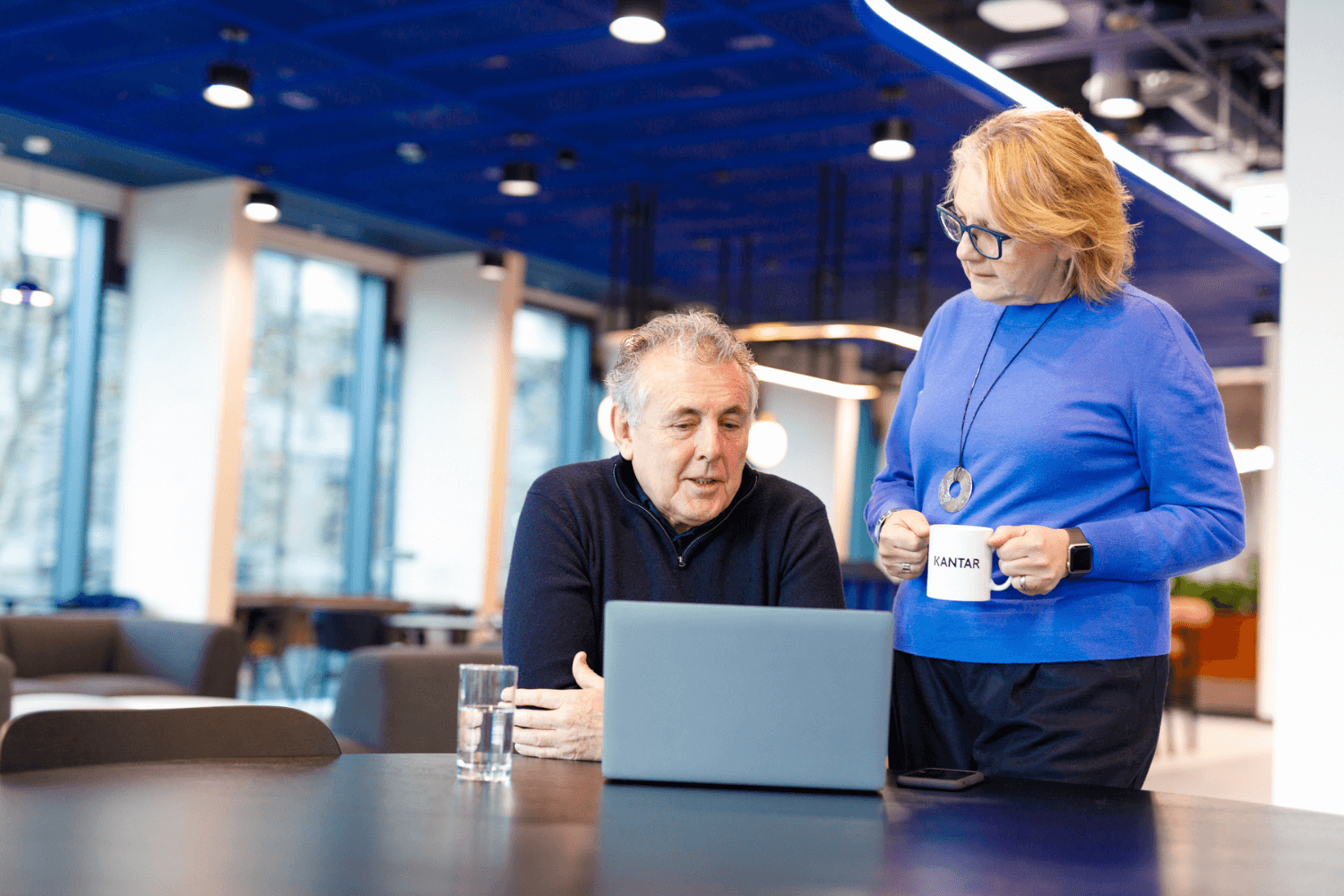 en vrouw in een blauwe trui staat rechtop en houdt een koffie vast. Aan tafel zit een man in een zwarte trui met zijn armen over elkaar, kijkend naar een laptop.