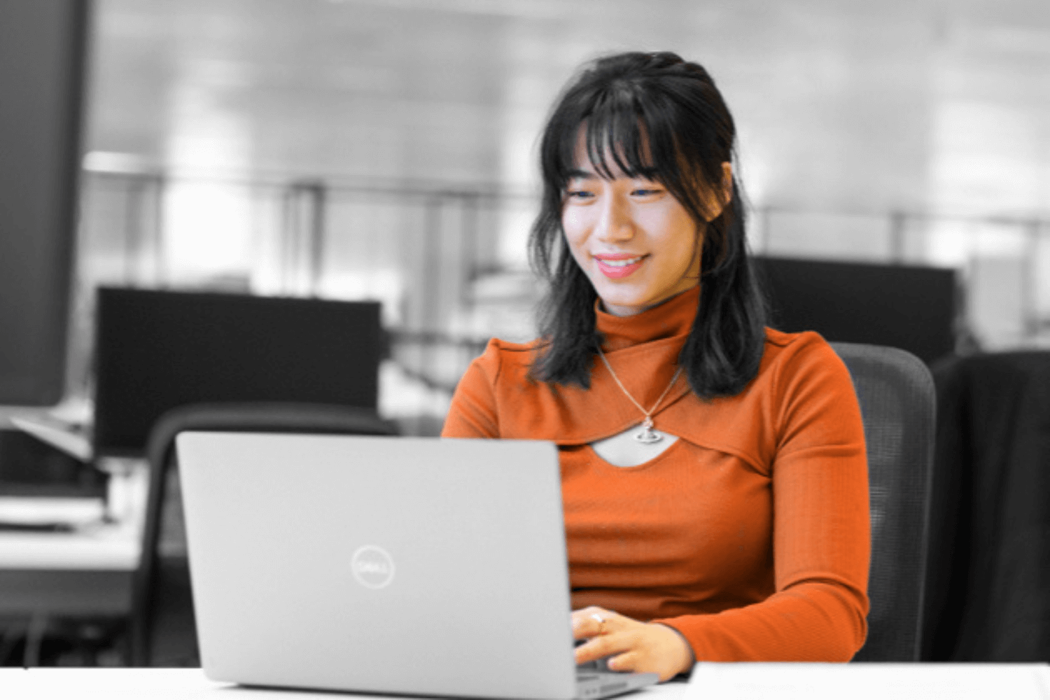 Zwart-wit afbeelding, waarbij alleen kleding en huidskleur in kleur zijn. Een jonge vrouw in een oranje trui werkt op een laptop.