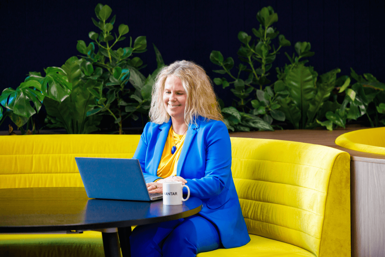 一位留着金色长卷发的女士坐在黄色的沙发上操作笔记本电脑。