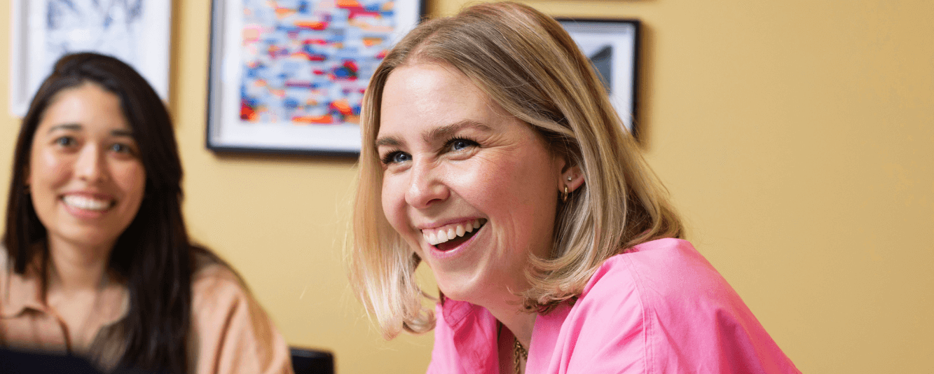 一个短发金发女人穿着粉红色的衬衫，对着镜头笑。她坐在一个涂成亮黄色的房间里。她身后是另一位女性同事，稍微有些模糊。她有长长的棕色头发，穿着棕色的上衣。