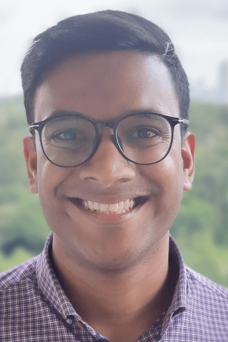 一位戴着圆形黑边眼镜的年轻人对着镜头微笑。他穿着格子衬衫，留着棕色短发。