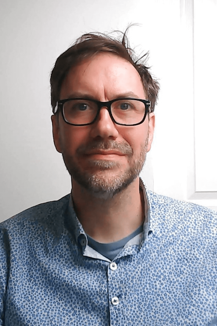 一名男子戴着黑色边框眼镜，身穿蓝色衬衫，正看着镜头。他有一头棕色短发。